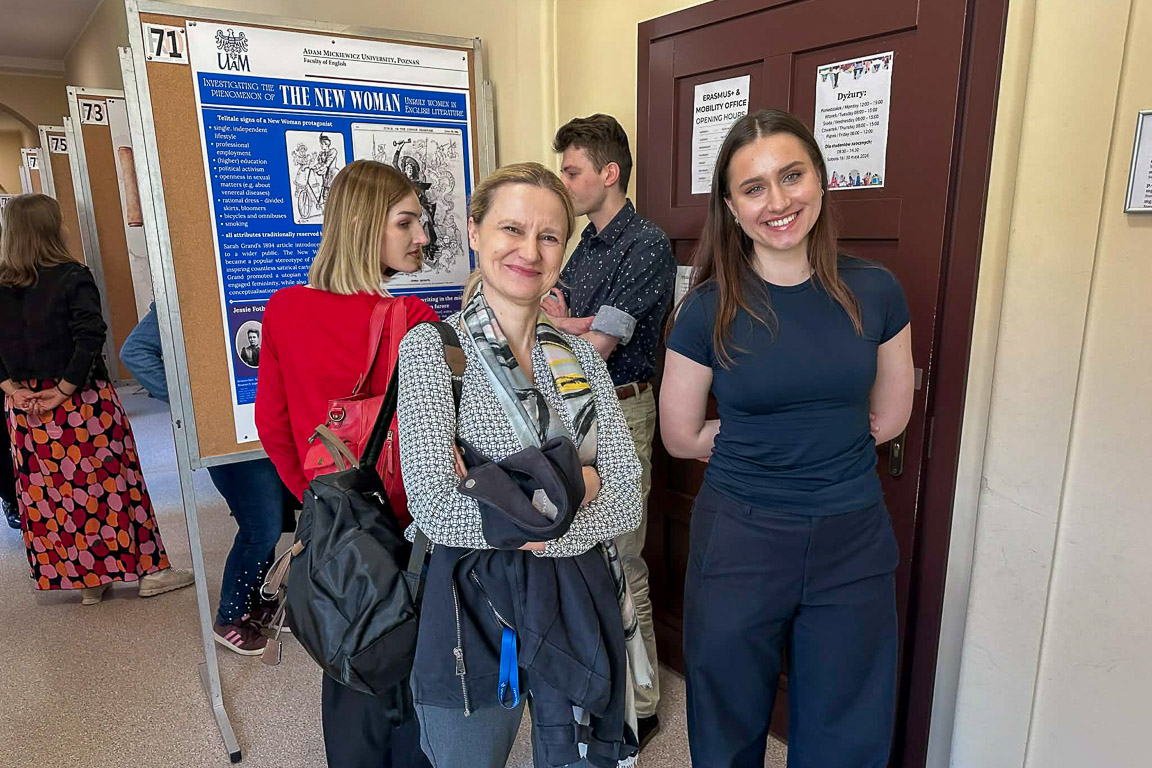 photograph: two persons posing in front of a conference poster stand; in the background: two other people looking at the poster