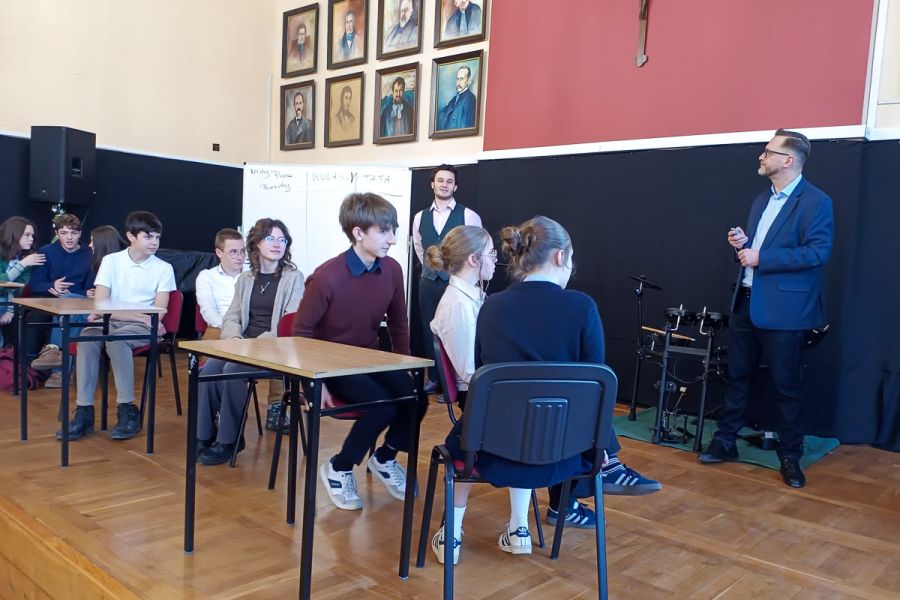 zdjęcie: dwie osoby stoją, dziesięcioro siedzi przy stołach w sali