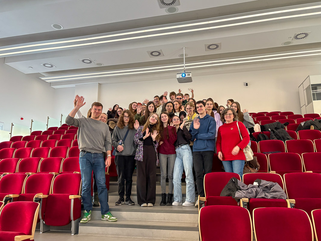 Grupa uczestników stoi na schodach w auli, uśmiecha się i macha w stronę aparatu, otoczona rzędami czerwonych krzeseł.