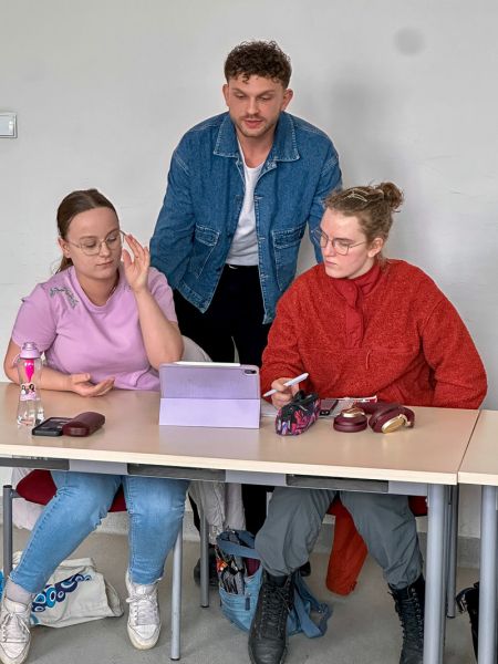 zdjęcie: dwie osoby siedzą za stołem, trzecia osoba stoi za nimi; rozmawiają