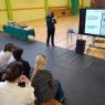 zdjęcie: sala gimnastyczna; osoba stoi, wskazując na slajd z prezentacji multimedialnej wyświetlony na ekranie; grupa osob siedzi na ławach i przygląda się prezentacji