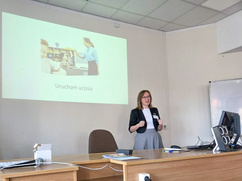 photograph: a person stands behind a table in a seminar room; a slide from a multimedia presentation is displayed on a screen behind them