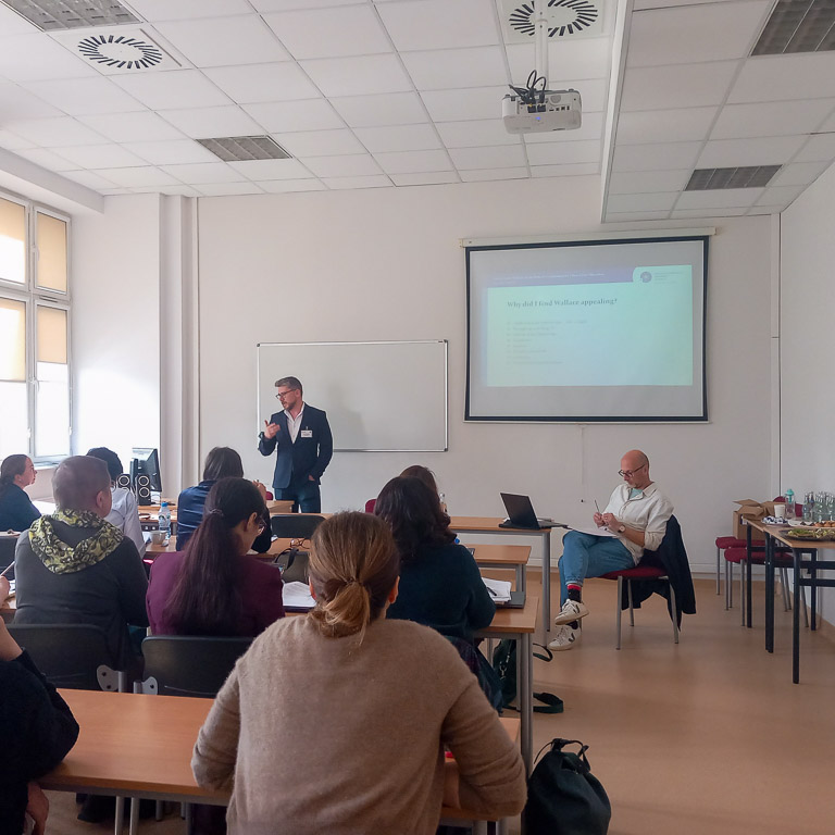 photograph: a person stands behind a table in the seminar room; a multimedia presentation slide is displayed on a screen behind them; the audience is gathered in the room in front of them