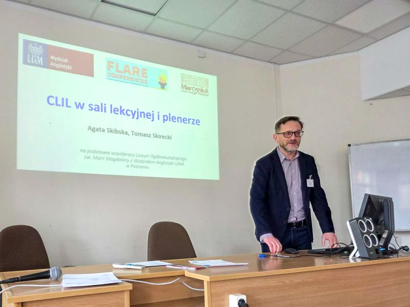zdjęcie: osoba stoi za stołem w sali seminaryjnej; za nią na ekranie slajd z prezentacji multimedialnej