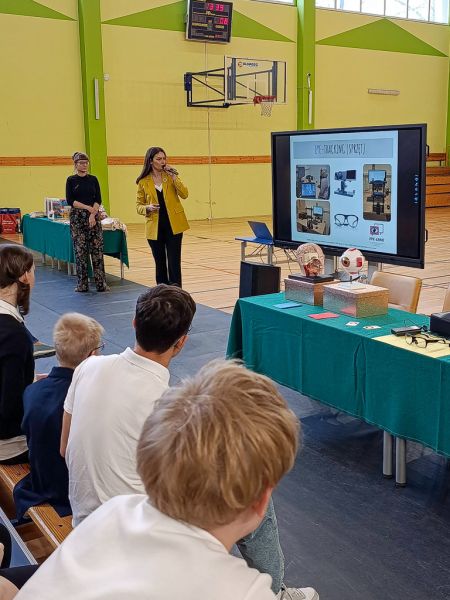 zdjęcie: sala gimnastyczna; dwie osoby stoją, jedna patrzy na slajd z prezentacji multimedialnej wyświetlony na ekranie; grupa osob siedzi na ławach i przygląda się prezentacji