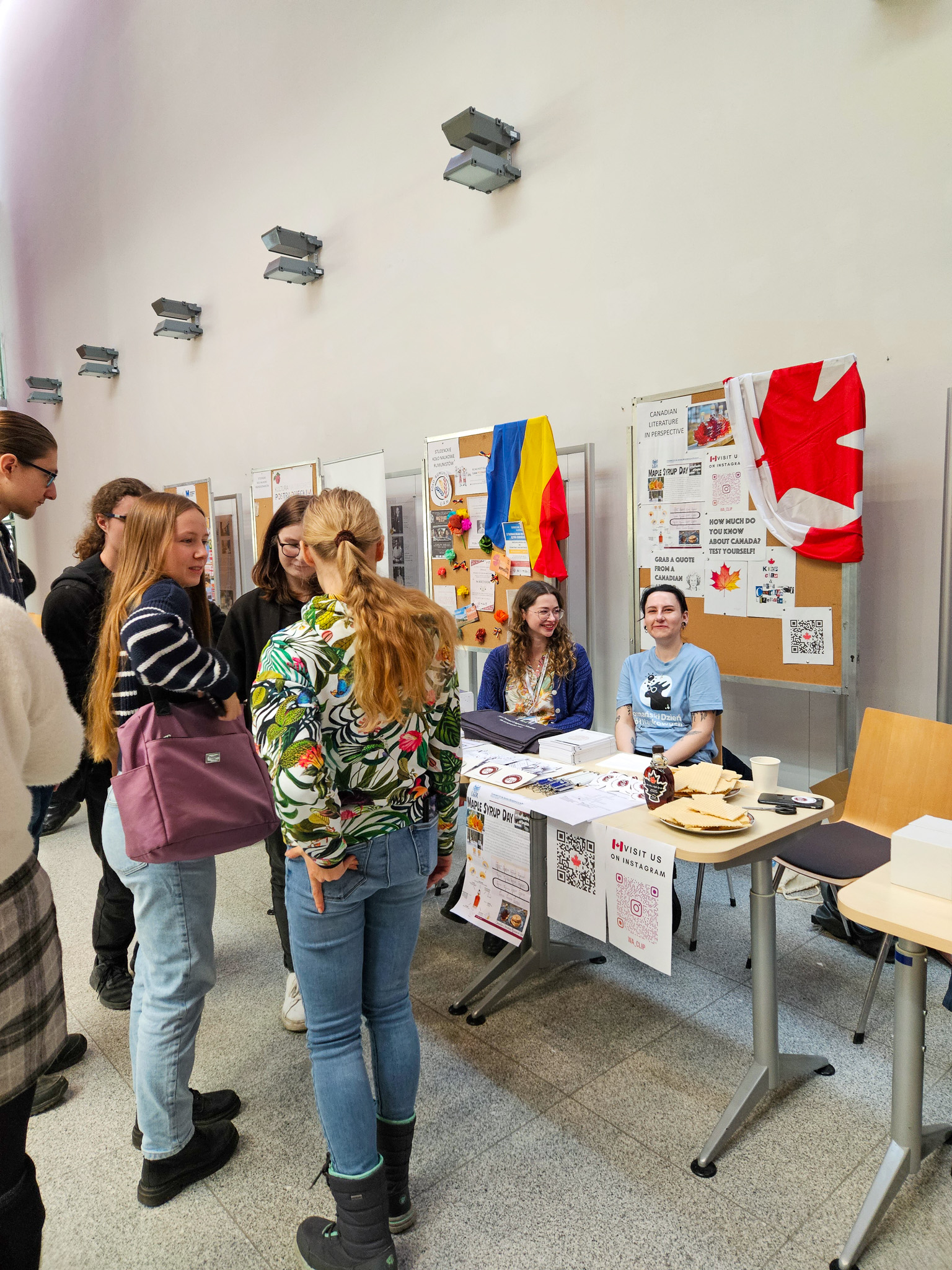 fotografia ilustrująca stoisko koła naukowego Canadian Literature in Perspective: dwie osoby siedzące za stołem, spoglądające w stronę obiektywu, oraz sześcioro osób stojących w pobliżu tego stołu