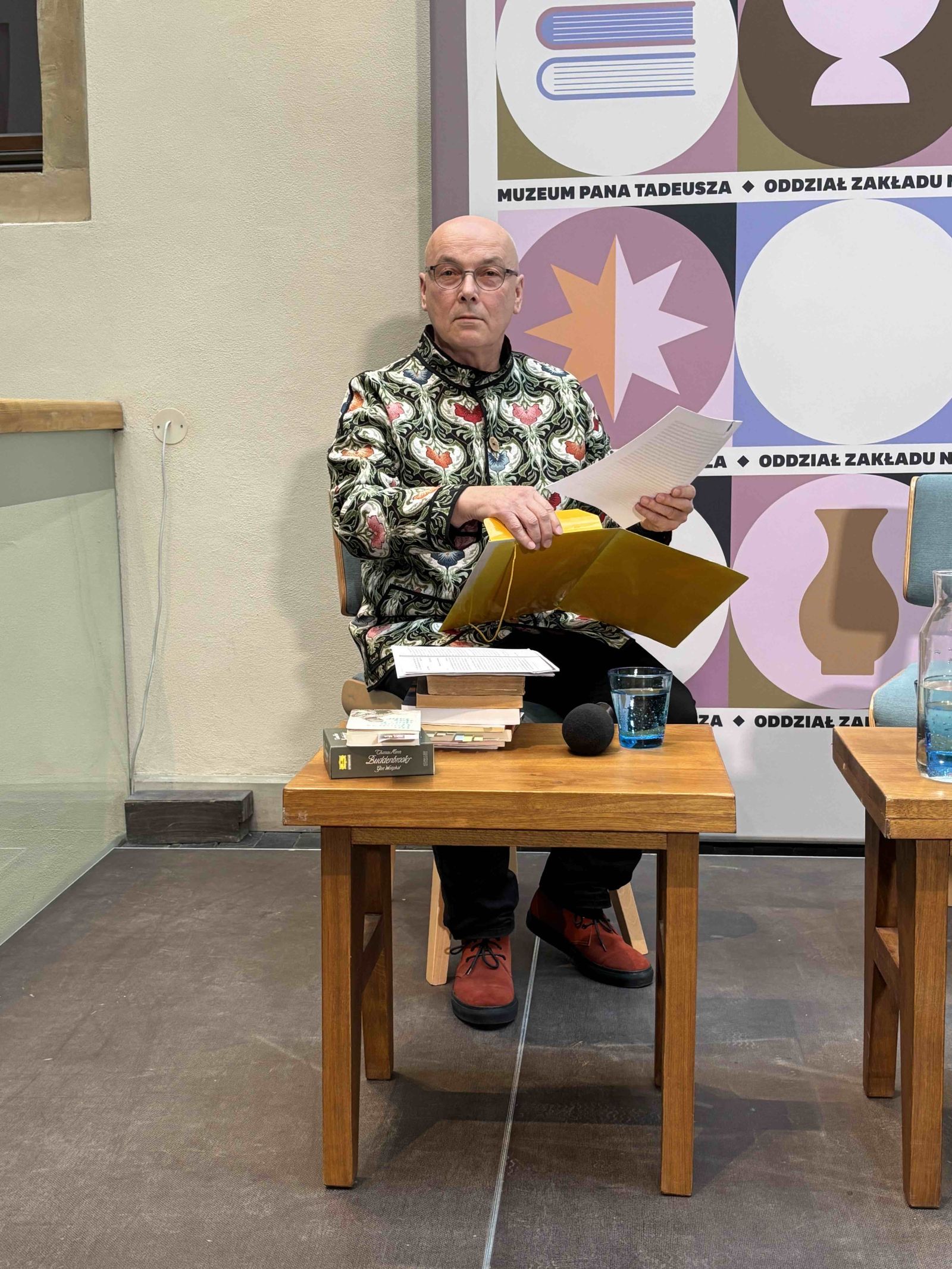 Zdjęcie przedstawiające siedzącego przy stoliku prof. Jerzego Kocha wyjmującego kartki z żółtej teczki