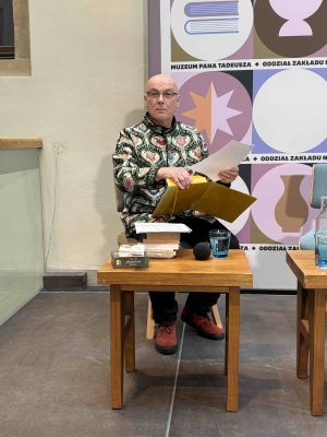 Zdjęcie przedstawiające siedzącego przy stoliku prof. Jerzego Kocha wyjmującego kartki z żółtej teczki