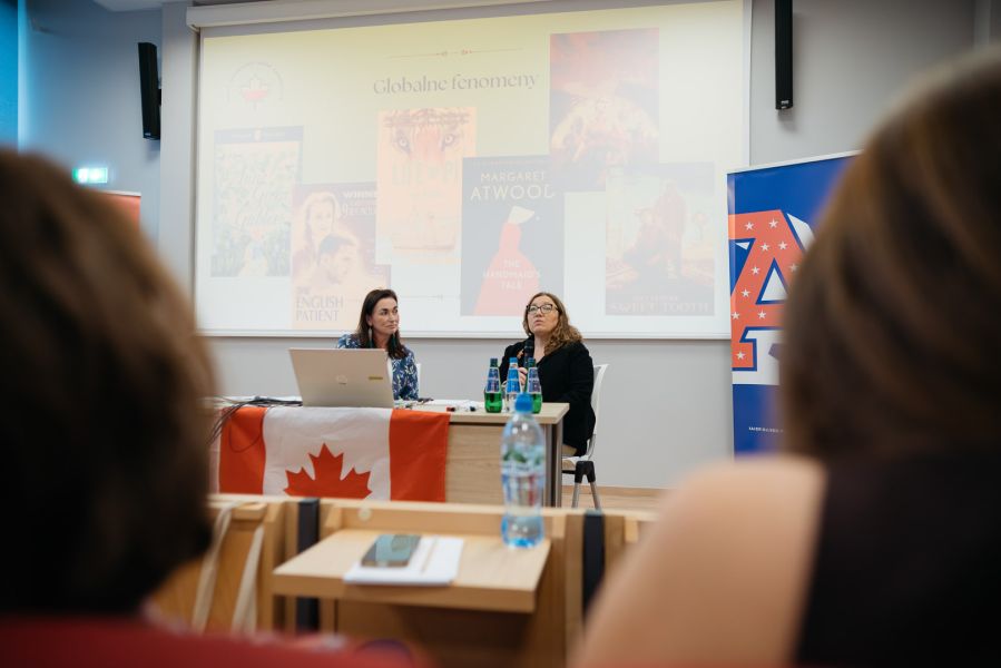 zdjęcie: dwie osoby siedzą za stołem w sali seminaryjnej; na ekranie za nimi: sjald z prezentacji; w sali przed nimi są widoczne nieostre sylwetki dwóch osób tyłem do obiektywu