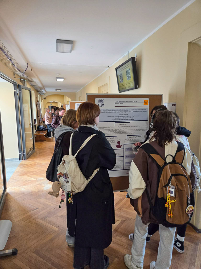zdjęcie: cztery osoby stoją przy tak zwanym posterze konferencyjnym rozmawiając; w tle kilka osób przy innych posterach ustawionych w korytarzu budynku