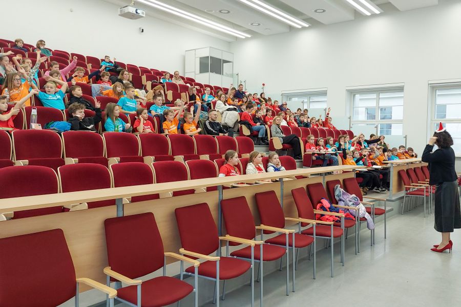 zdjęcie: sala wykładowa: wykładowca stoi, grupa kilkudziesięciu dzieci oraz ich opekunowie siedzą na sali i słuchają wykładu