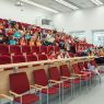 zdjęcie: sala wykładowa: wykładowca stoi, grupa kilkudziesięciu dzieci oraz ich opekunowie siedzą na sali i słuchają wykładu