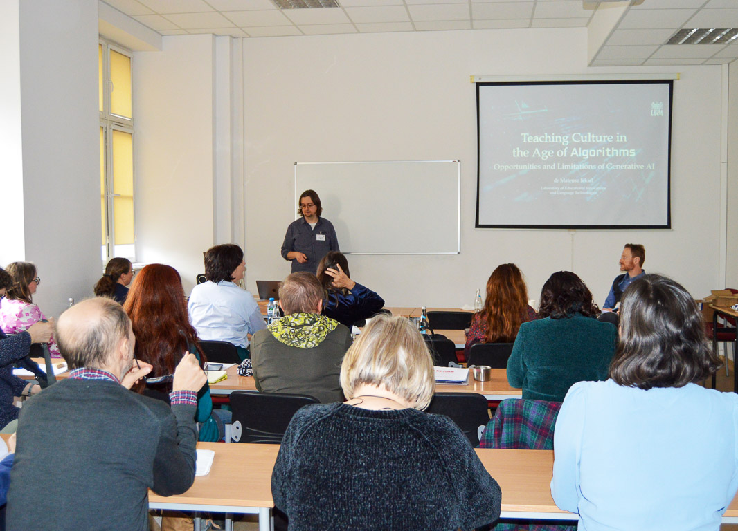 photograph: a person stands behind a table in the seminar room; a multimedia presentation slide is displayed on a screen behind them; the audience is gathered in the room in front of them