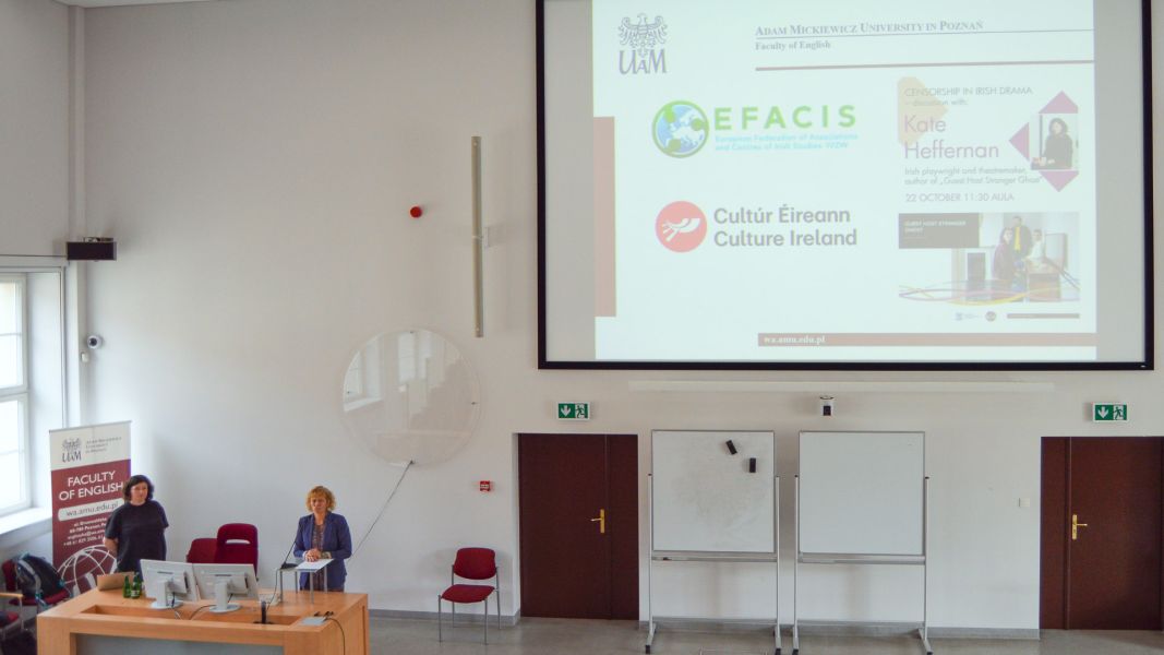 photograph: a person addresses the audience from behind the pulpit; another person looks at the audience (not visible in the frame); also in the frame: a screen with the title card of the presentation