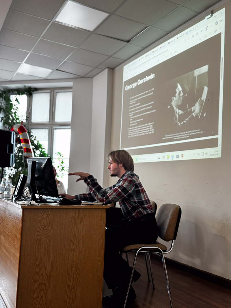 zdjęcie: osoba siedzi w sali seminaryjnej; w tle, na ekranie, slajd z prezentacją multimedialną