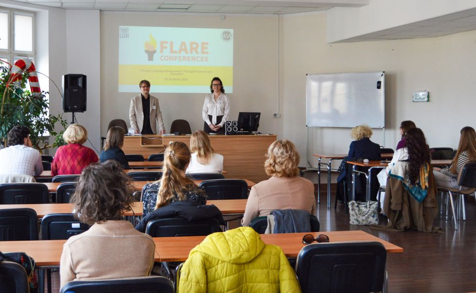 zdjęcie: dwie osoby stoją za stołem w sali seminaryjnej; za nimi na ekranie slajd z prezentacji multimedialnej; przed nimi zgromadzona w sali publiczność