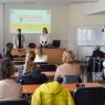 zdjęcie: dwie osoby stoją za stołem w sali seminaryjnej; za nimi na ekranie slajd z prezentacji multimedialnej; przed nimi zgromadzona w sali publiczność