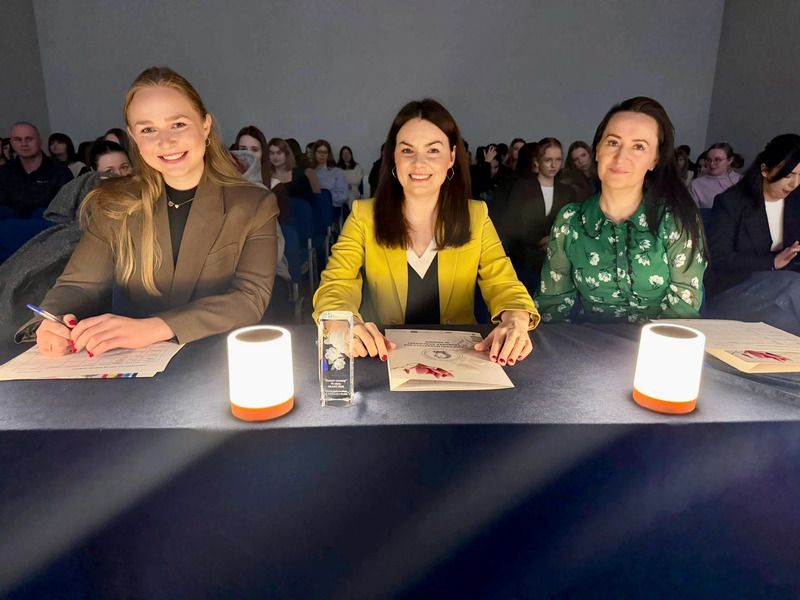 zdjęcie: trzy osoby stanowiące jury konkursu siedzą za stołem; za nimi, w tle, grupa kilkudziesięciu osób