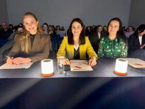 zdjęcie: trzy osoby stanowiące jury konkursu siedzą za stołem; za nimi, w tle, grupa kilkudziesięciu osób