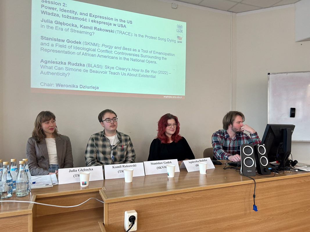 zdjęcie: cztery osoby siedzą za stołem w sali seminaryjnej; w tle, na ekranie, slajd z prezentacją multimedialną