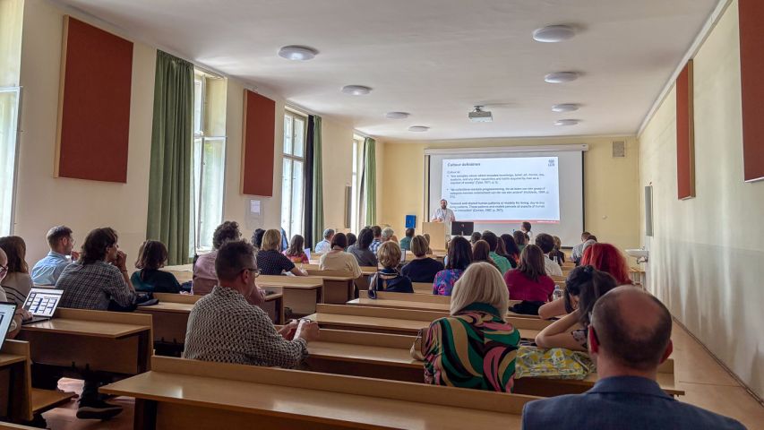 Widok sali wykładowej z publicznością siedzącą w ławkach; na froncie osoba przy mównicy prezentuje slajd z tekstem i schematem, widoczne wysokie okna po lewej oraz projektor pod sufitem.