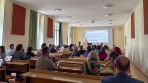 Widok sali wykładowej z publicznością siedzącą w ławkach; na froncie osoba przy mównicy prezentuje slajd z tekstem i schematem, widoczne wysokie okna po lewej oraz projektor pod sufitem.