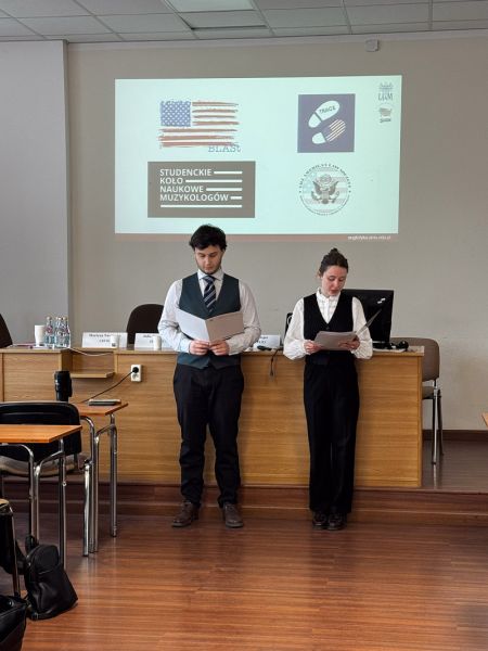 zdjęcie: dwie osoby stoją w sali seminaryjnej; w tle, na ekranie, slajd ze znakami biorących w wydarzeniu kół studenckich